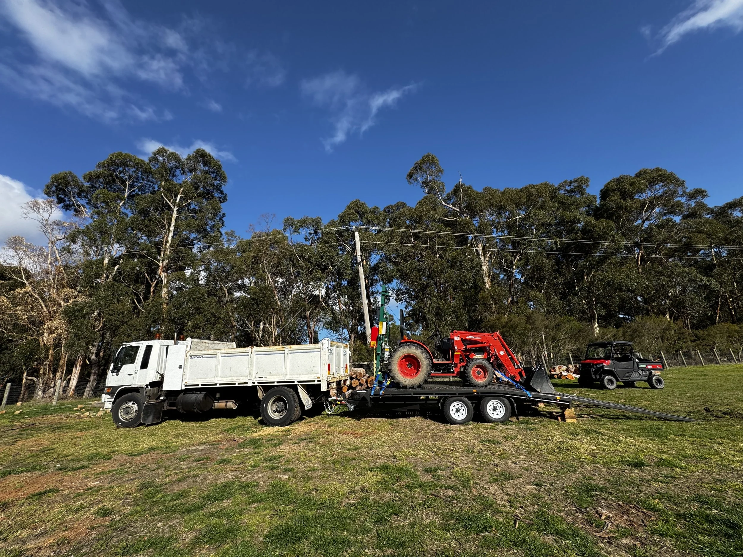 A white truck with a flatbed trailer carrying a red tractor and a black utility vehicle parked on a grassy field with trees and a blue sky in the background.