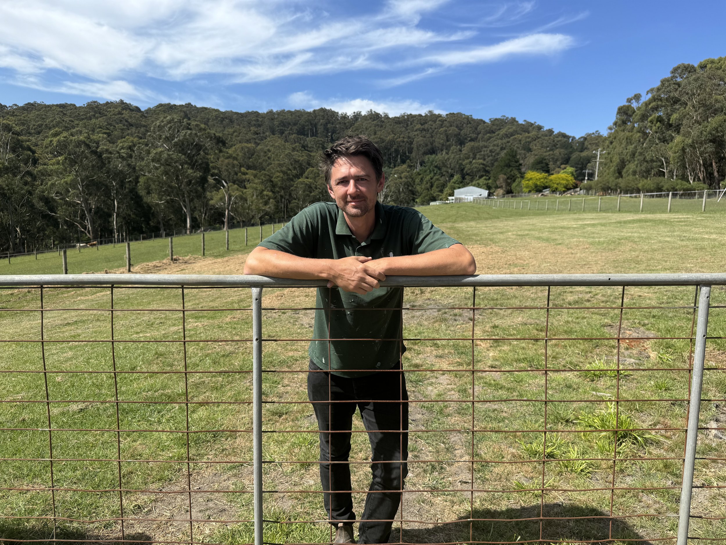A man leaning on a metal fence in a green field with trees and hills in the background under a partly cloudy blue sky.