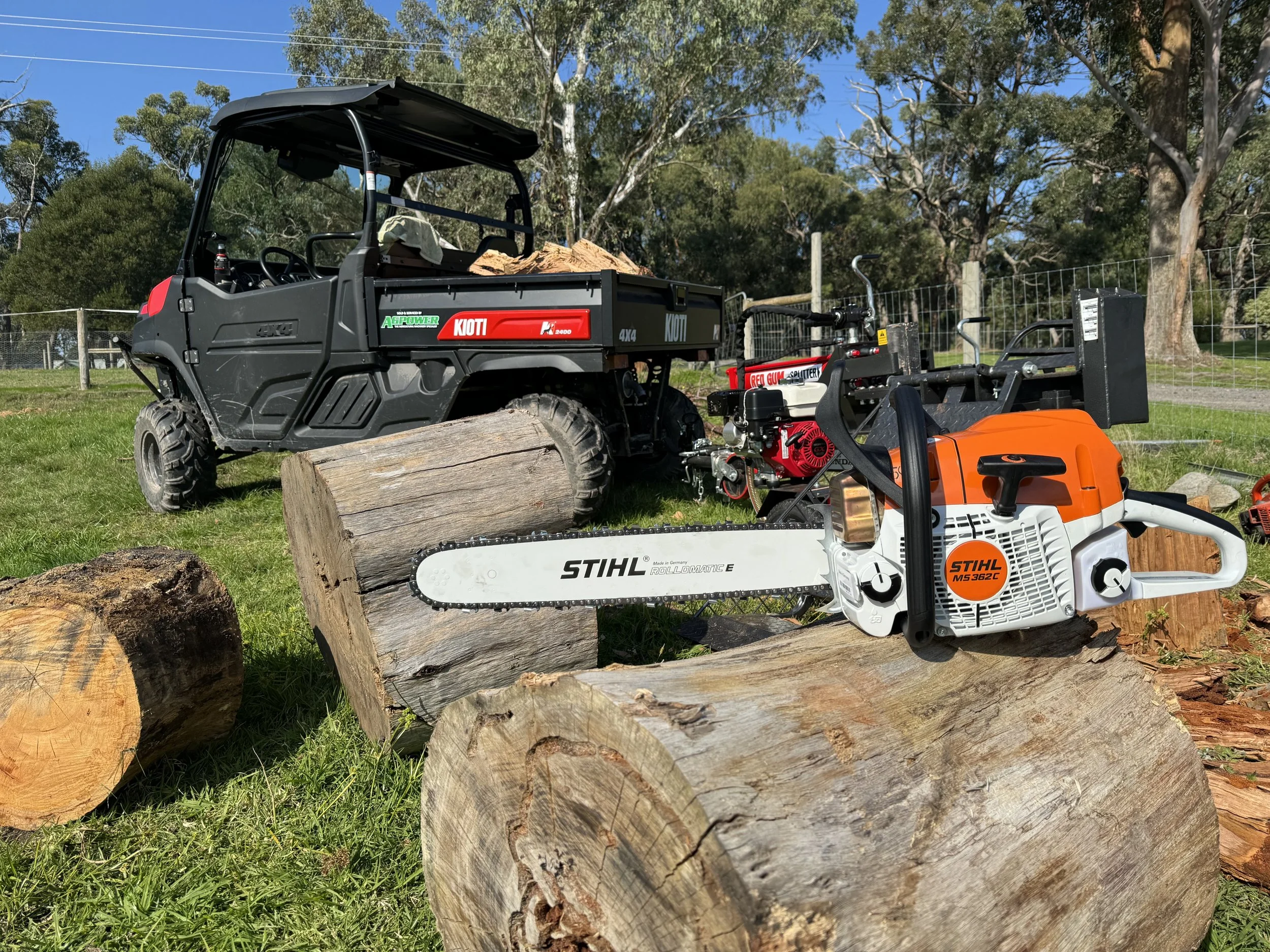 A chainsaw resting on a log with a utility vehicle and log splitter in the background in a grassy area surrounded by trees.