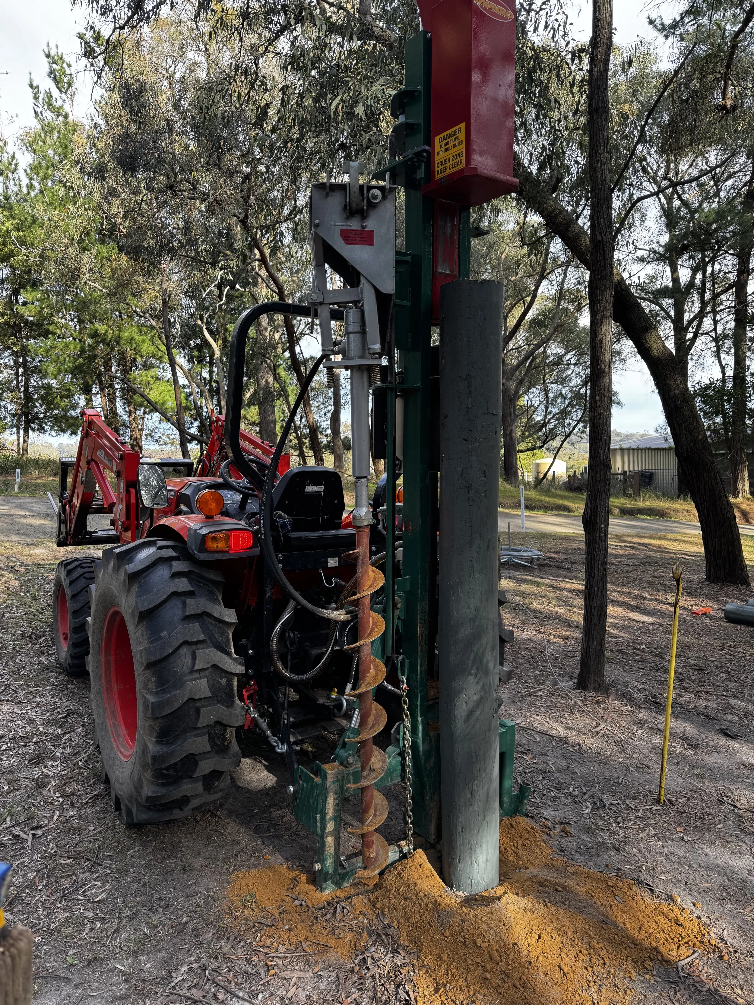 A piece of construction equipment with a large auger drill bit attached, positioned vertically on a dirt site surrounded by trees and a red tractor, with warning signs visible on the equipment.
