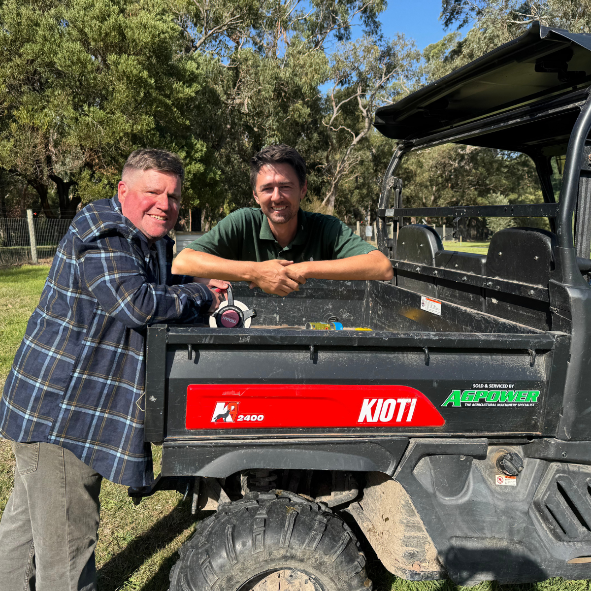 Two men smiling and leaning on a utility vehicle outdoors in a grassy area with trees in the background.