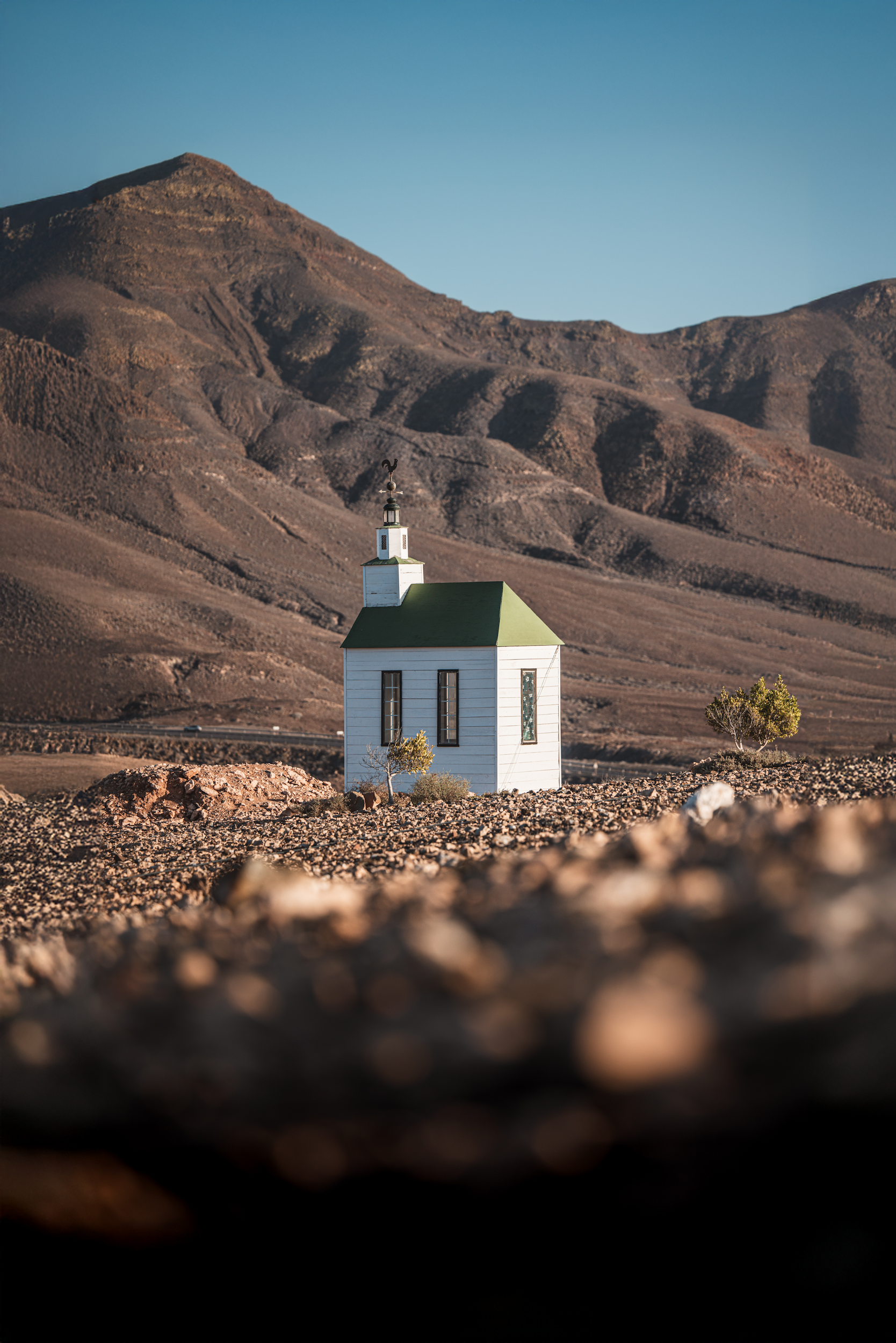 Small white church with green roof in rocky desert landscape with mountains in the background