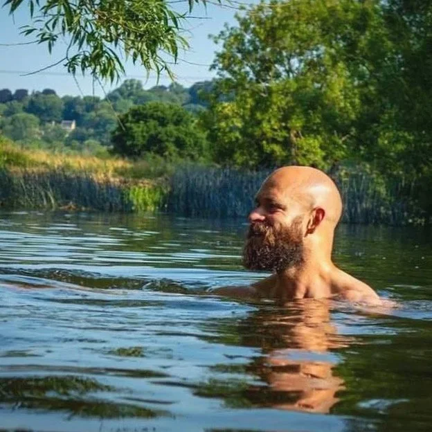 A bald man with a beard swimming in a calm river, surrounded by green trees and clear skies.