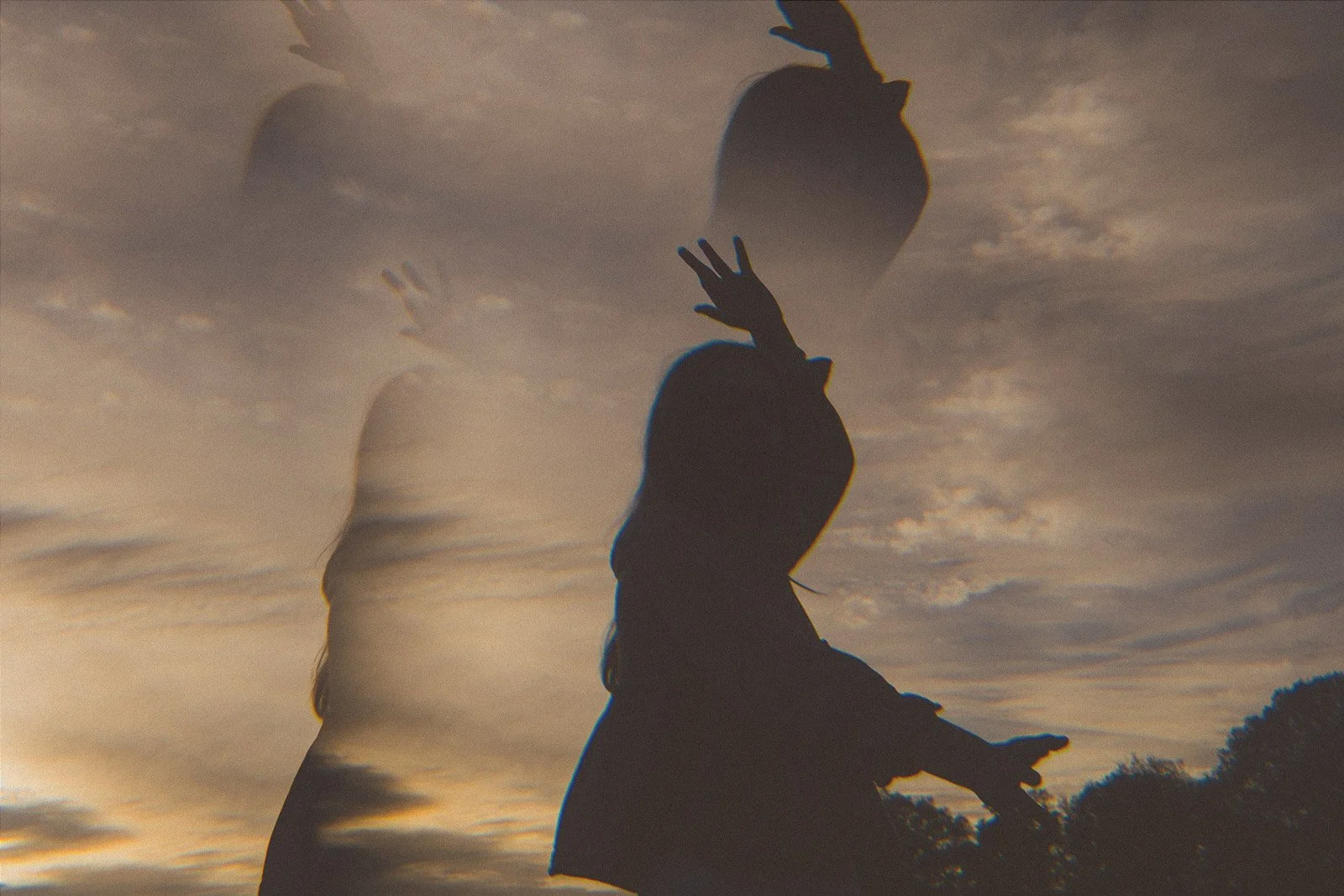 A double exposure photograph of a silhouetted woman, dancing and  reaching upward with both hands against a cloudy sky at sunset.