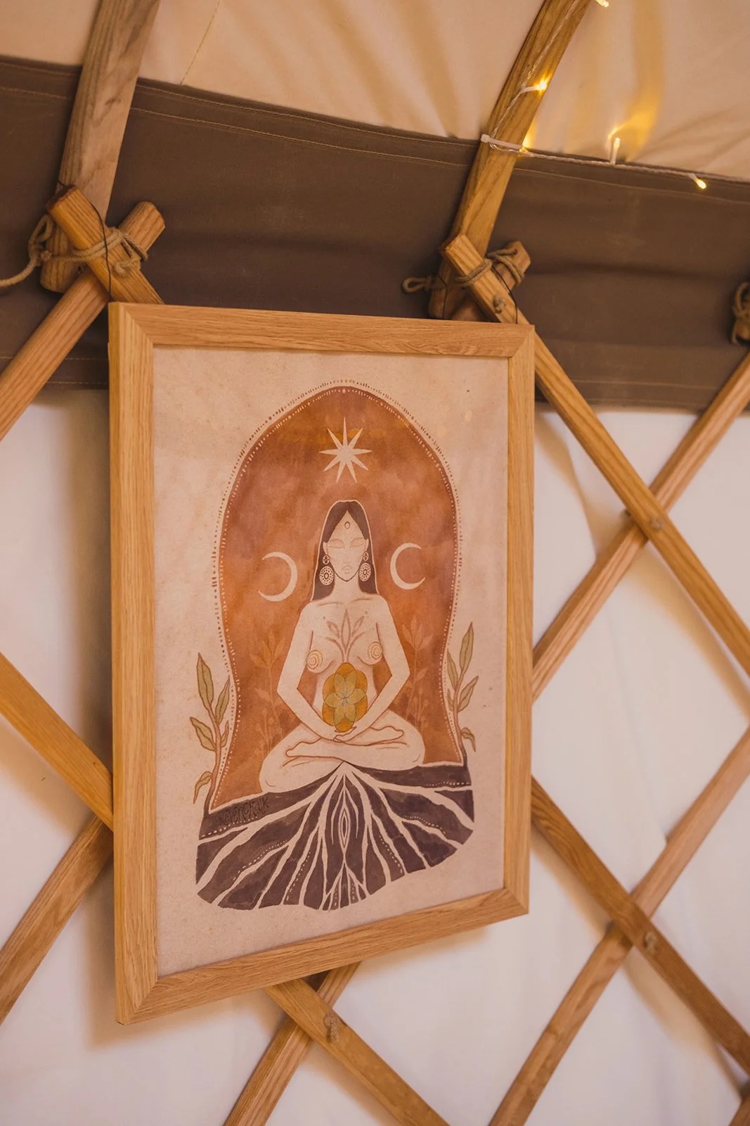 A framed artwork hangs on a wooden lattice inside a yurt with string lights. The artwork depicts a meditative woman with symbols of moon phases, stars, and roots.