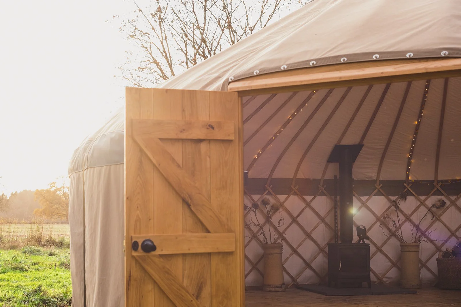 Open yurt with wooden door in natural surroundings, interior with wood stove and string lights, outdoor grass and trees visible, during daytime.