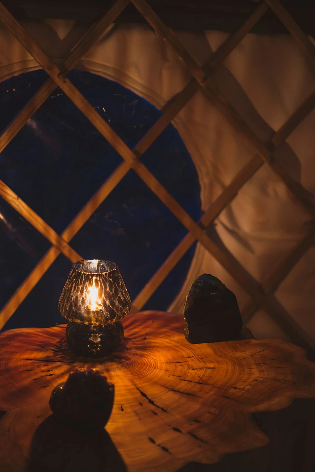 A cozy interior scene featuring a small table with a lit candle in a textured glass holder and a dark rock, set in front of a large window with a diamond-shaped grid. Outside the window, the night sky is visible.