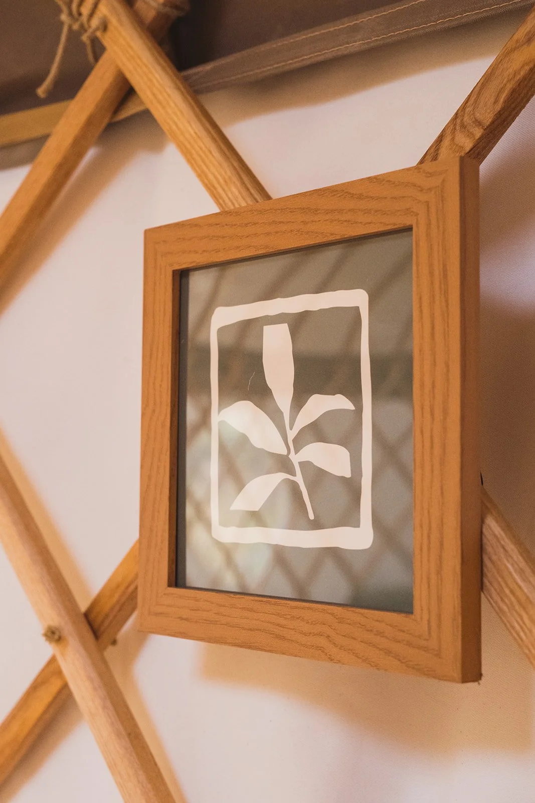 Close-up of a wooden framed mirror with a white leaf design on the glass, leaning against a wall with a wooden lattice structure in the background.