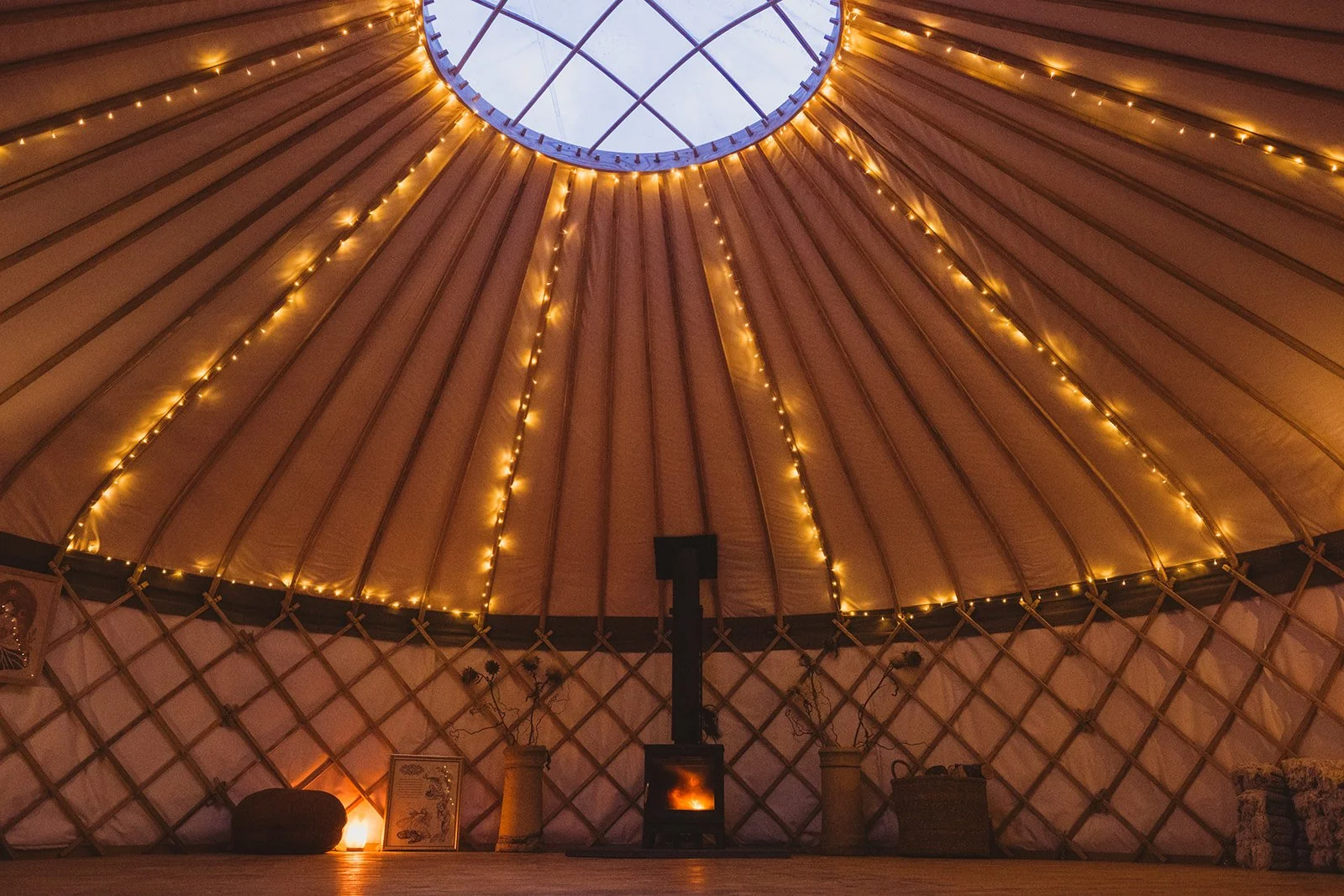 Interior of a yurt with beige fabric walls, a circular skylight at the top, and string lights hanging along the structure, with decorative vases, pictures, and a stove at the center.