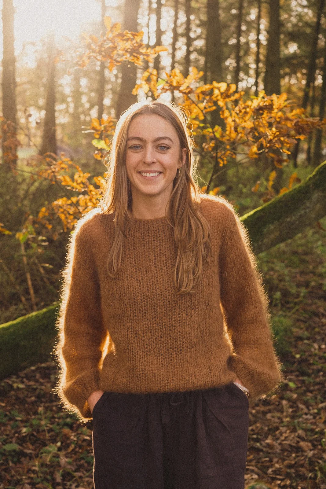 A woman, Hannah Marshall, smiling outdoors in a forest during autumn, wearing a brown sweater and black pants, with sunlight filtering through trees in the background.