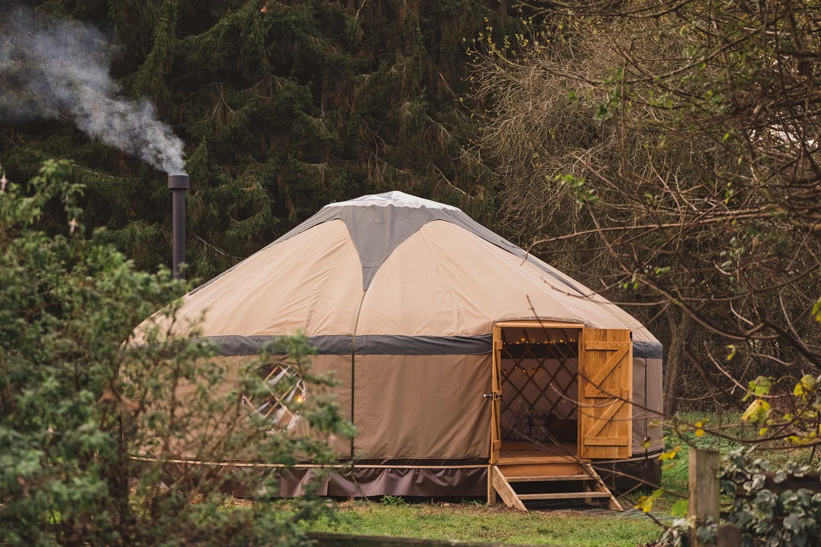 An inviting image of the exterior of The Whiteleaf Yurt amongst picturesque trees and vegetation, with the chimney gently smoking and the double doors open.