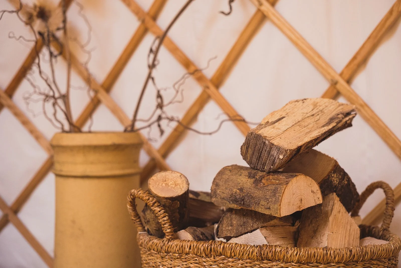 Wicker basket filled with chopped firewood logs next to a tall yellow vase with dried branches, set against a wooden lattice wall backdrop.