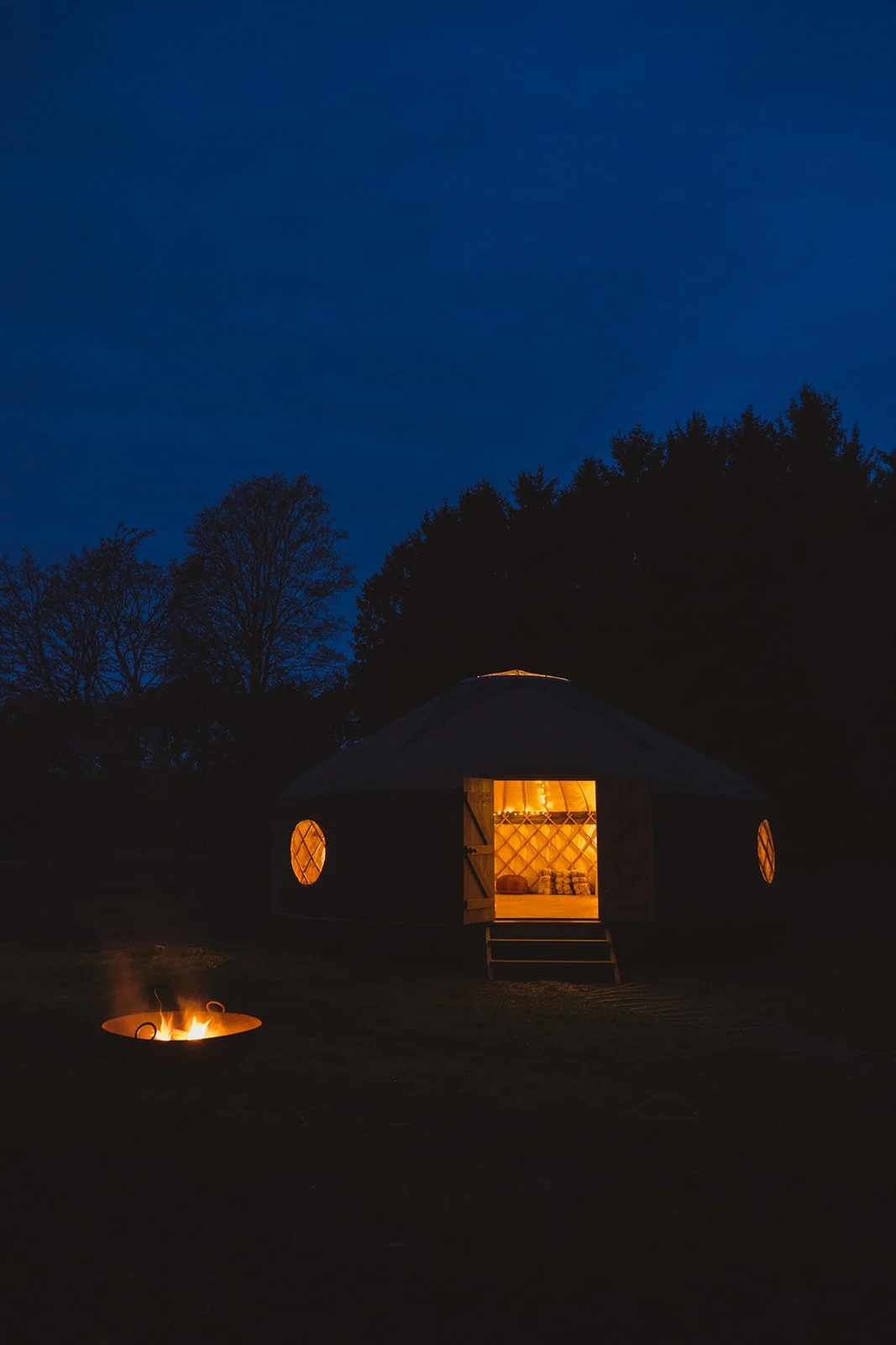 A yurt with open door illuminated from inside, sitting in a field at night with trees in the background; a fire pit with a small fire burning is in the foreground.