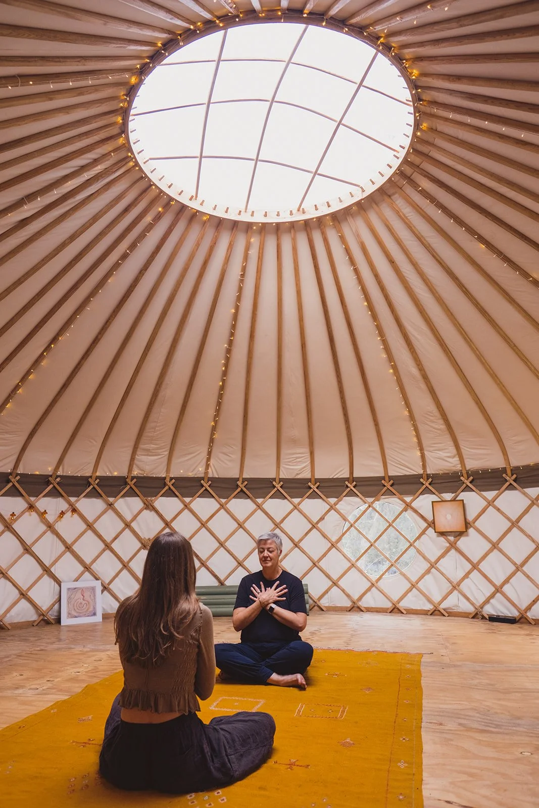 Two women seated on a yellow rug inside a round yurt with wooden framing, one practicing yoga or meditation with hands on chest, the other facing away; decorative art and a circular window are visible on the yurt wall.