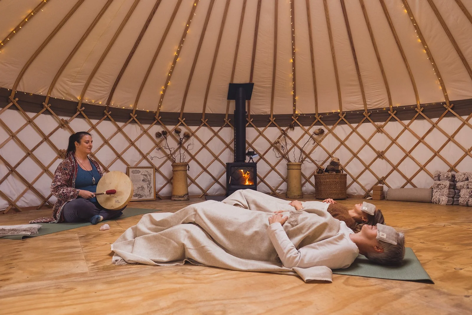 Two women lying on yoga mats inside a yurt, with a woman playing a shamanic drum, and a small fire burning in a wood stove at the center of the space.