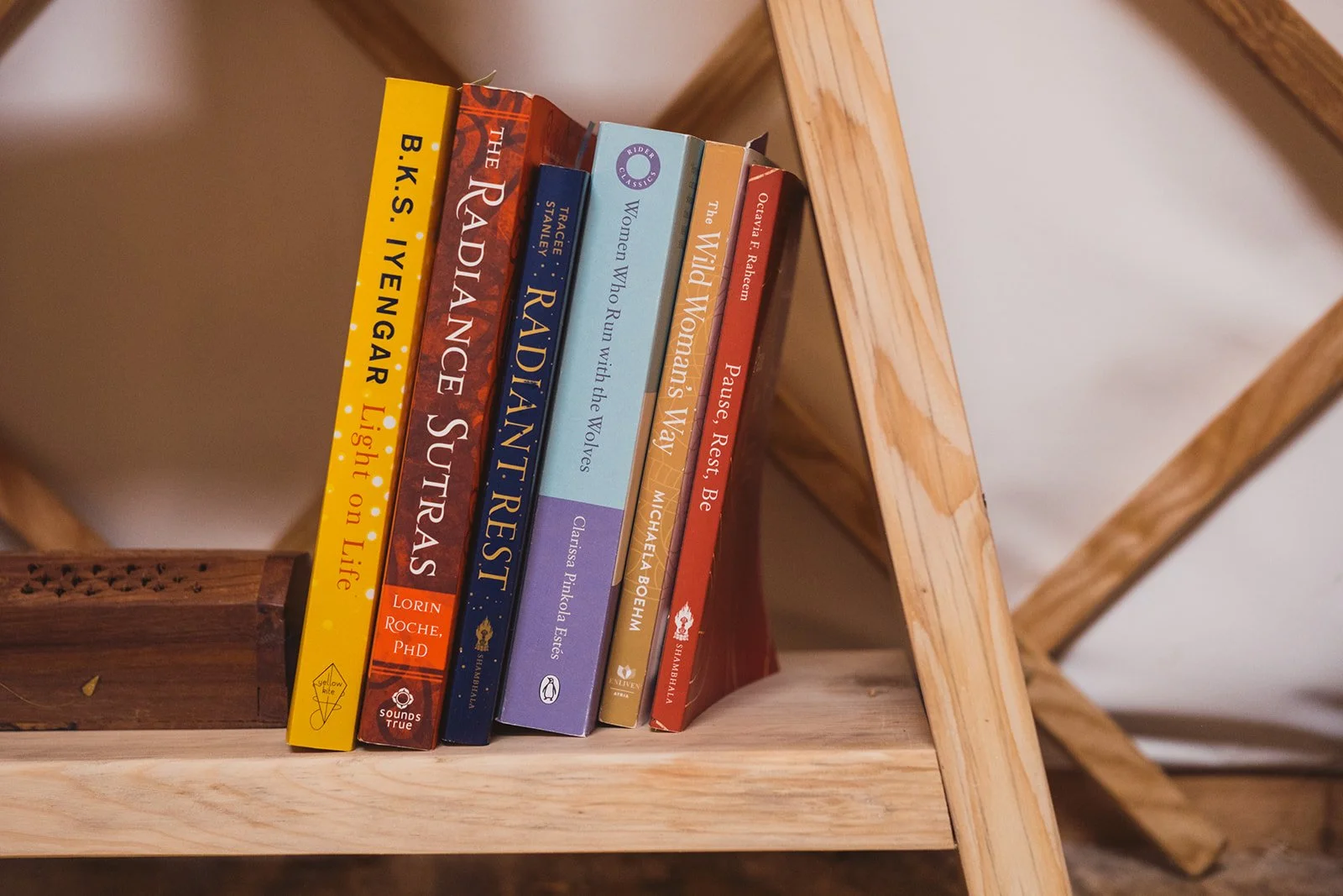 A row of seven books on a wooden shelf, with titles including 'B.K.S. Iyengar Light on Life,' 'The Radiance Sutras,' 'Raindance,' 'Women Who Run with the Wolves,' 'The Wild Women Way,' and 'Pause Rest Be.'