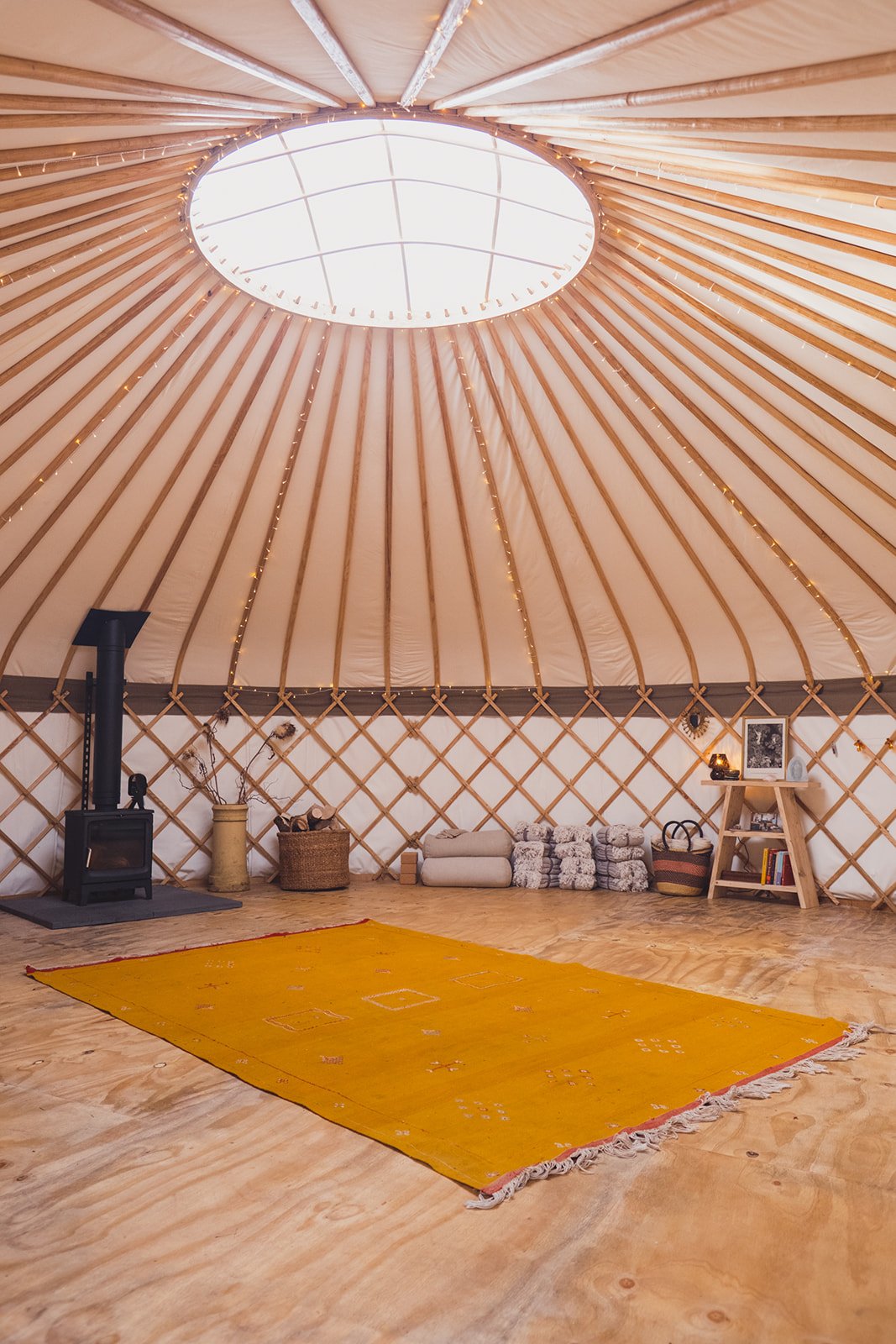 Interior view of The Whiteleaf Yurt, with a woodburner, a yellow carpet, and equipment neatly stacked.