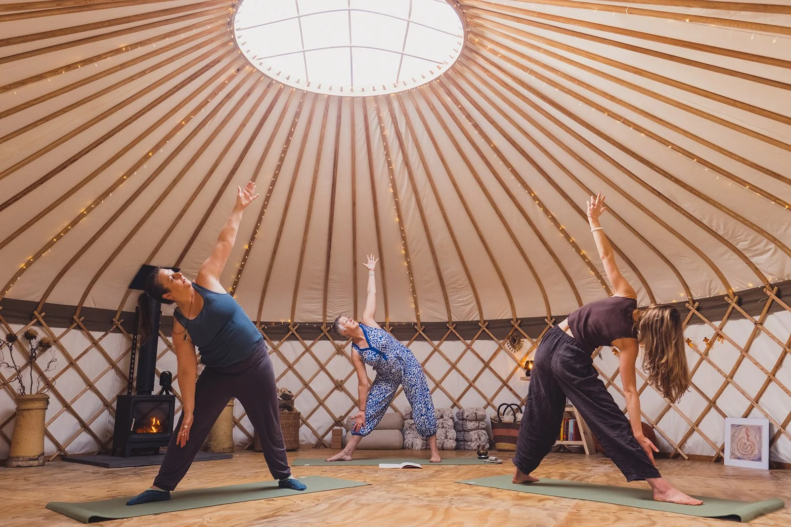 Three women practicing yoga inside a yurt with wooden walls and a circular skylight on the ceiling. They are in different yoga poses on yoga mats, with a cozy fireplace, books, and framed art in the background.