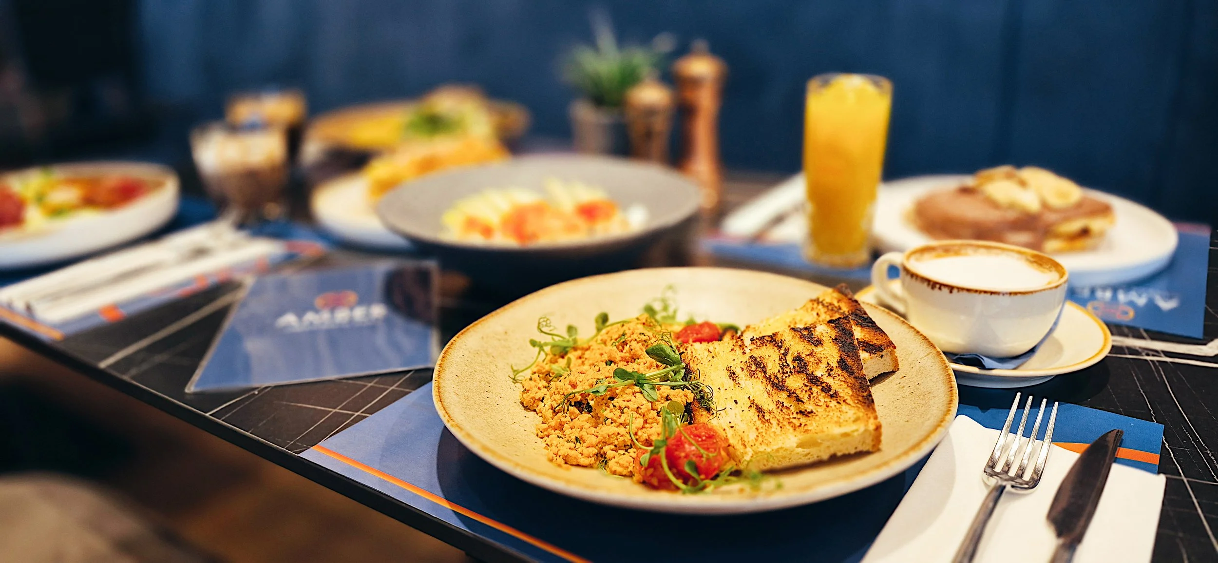 Breakfast plated with toast, scrambled eggs, garnished with herbs, and tomatoes, alongside a cup of coffee with foam, a glass of orange juice, and additional plates of food on a table.