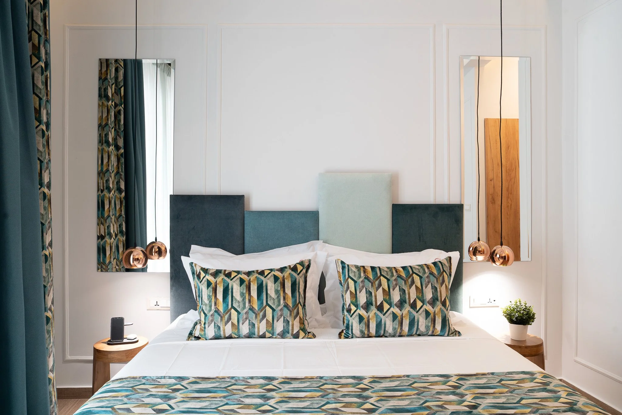 A neatly made bed with patterned pillows and a colorful bedspread, flanked by two wooden nightstands with lamps, a potted plant, and mirrors on the wall.