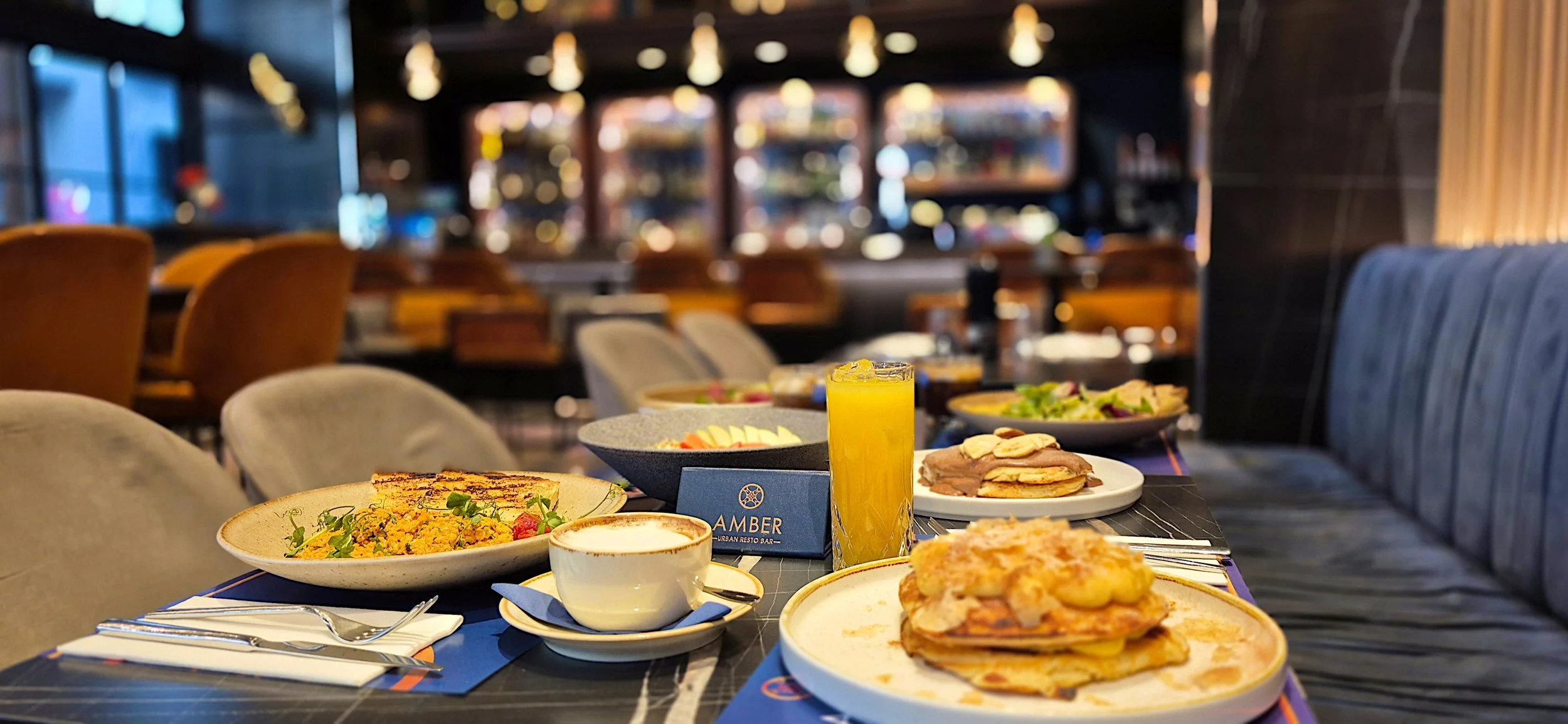 A table set with various breakfast dishes including pancakes with bananas and syrup, a bowl of salad, a plate of scrambled eggs, a cup of coffee, a glass of orange juice, and a business card that reads 'Amber'. The background shows a modern restauran