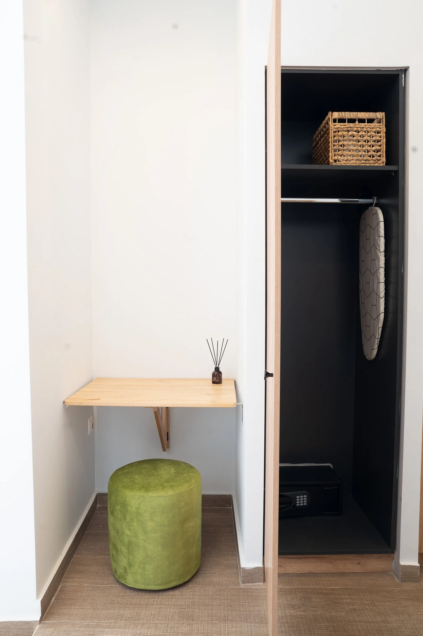 A small corner with a wooden desk, a round green ottoman, and an open closet with a safe, an ironing board, and a wicker basket.