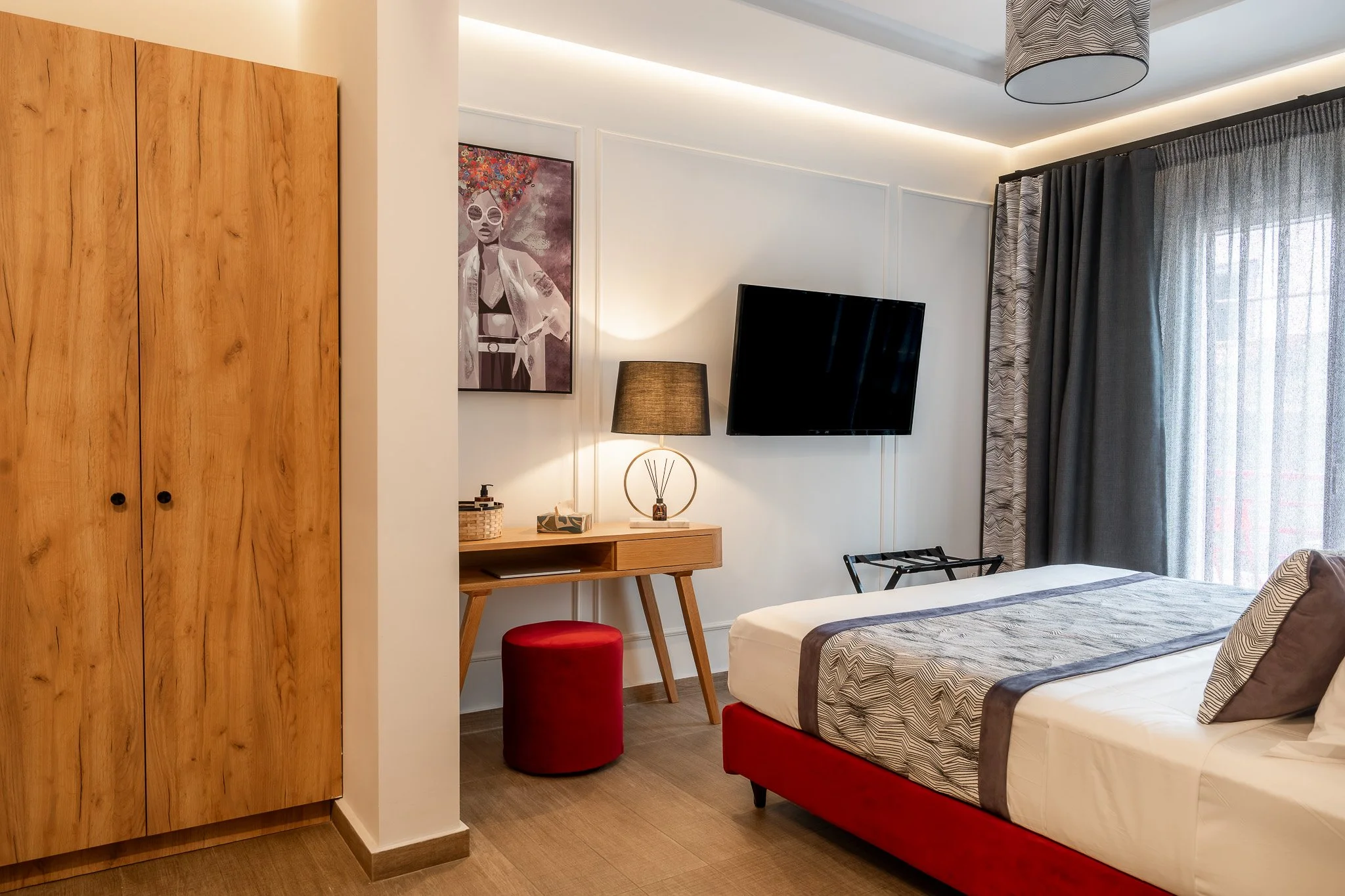 Modern hotel room with a bed, wooden wardrobe, wall-mounted TV, desk with a lamp, artwork, and curtains.