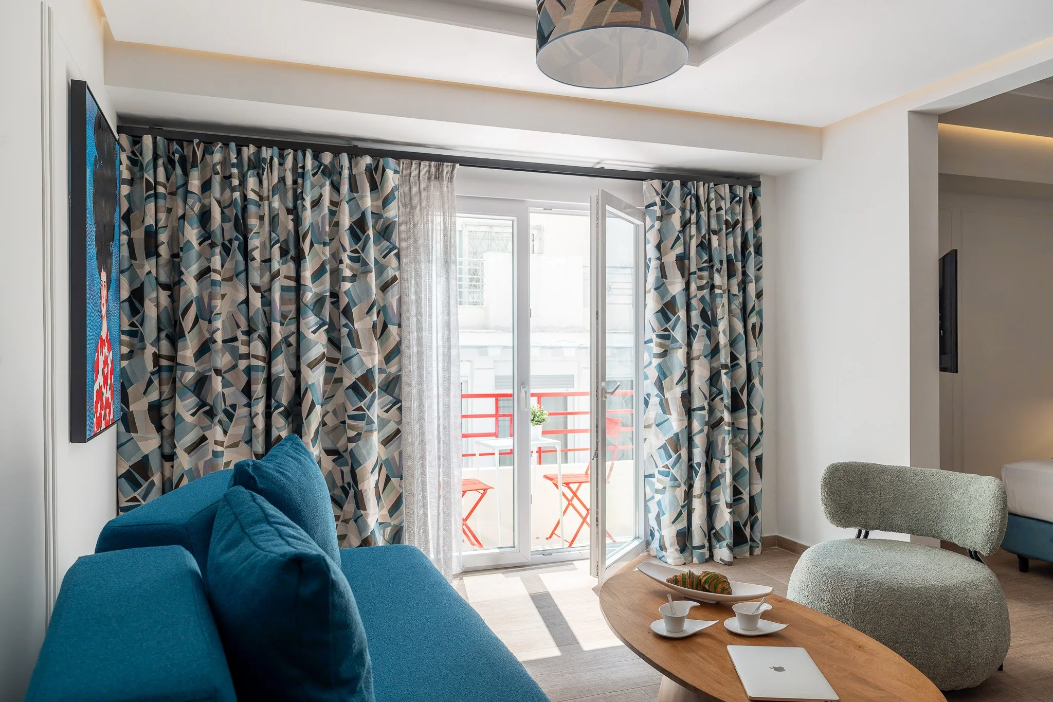 Living room with a sliding glass door opening to a balcony, patterned curtains, a blue sofa with cushions, a light green modern chair, a wooden coffee table with teacups and an open laptop, and a mounted flat screen TV.