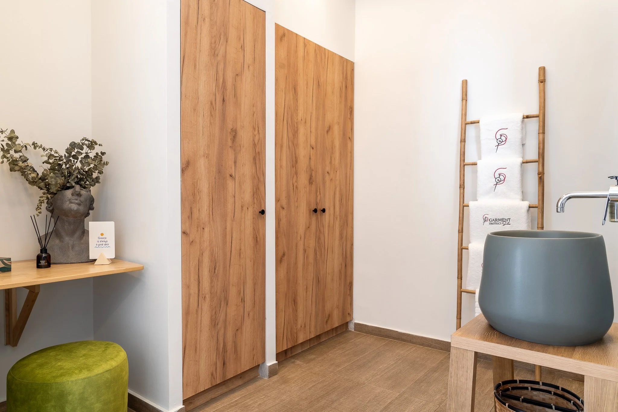 Interior of a room with wooden cabinets, a small table with decorative items, a green ottoman, a bamboo towel ladder with white towels, and a large gray planter on a wooden stand.