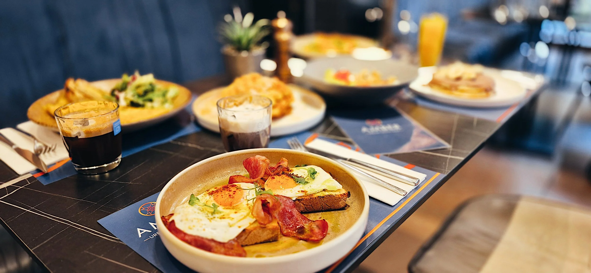 A breakfast table with plates of eggs, bacon, toast, salad, and drinks, set on a black table with blue placemats.