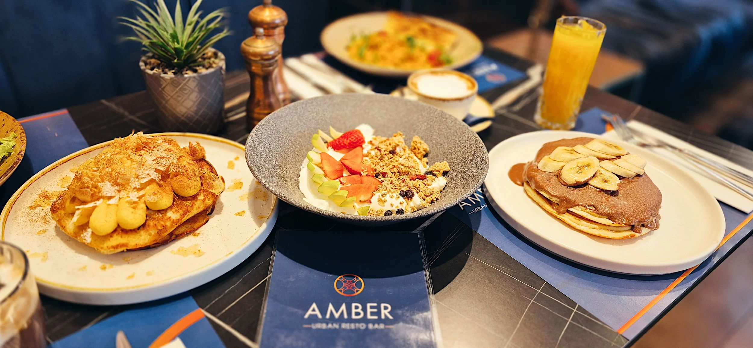 Spread of breakfast dishes on a table including a plate with a waffle topped with strawberries and bananas, a bowl with yogurt, granola, and strawberries, and a plate with pancakes topped with sliced bananas. There is also a glass of orange juice, a 