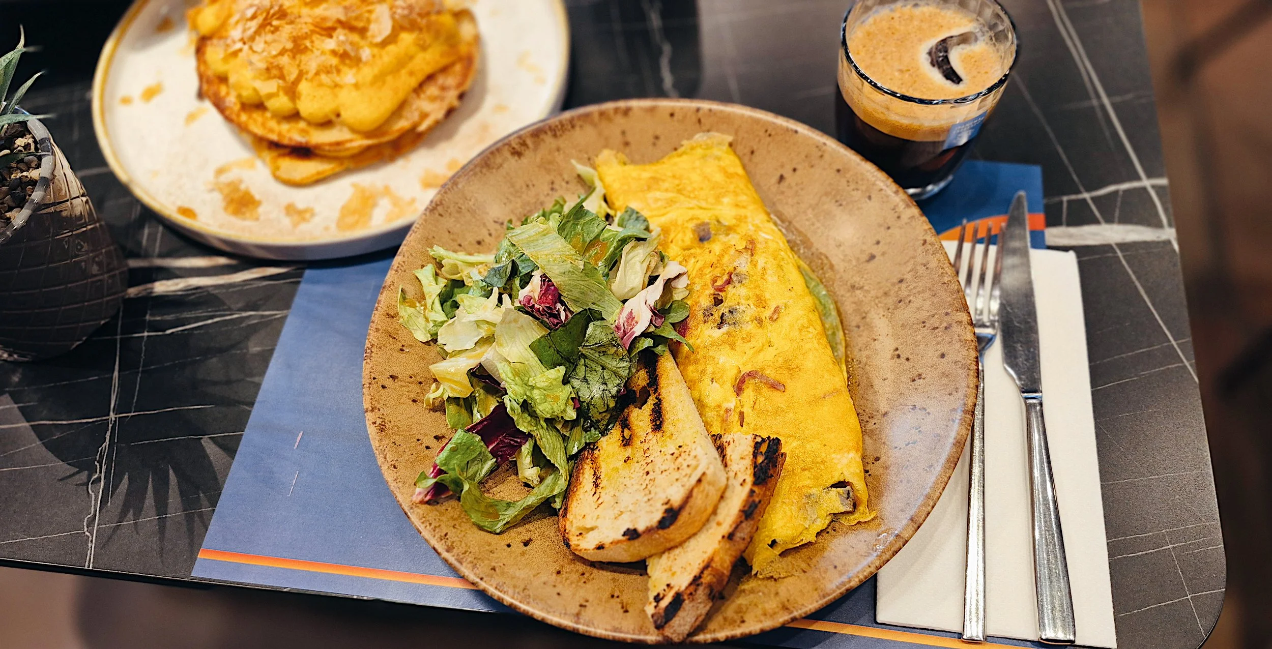 Plate with salad, grilled toast, and a folded omelet on a table set with a fork, knife, and napkin. In the background, a side plate with a biscuit or bread and a glass of dark soda.