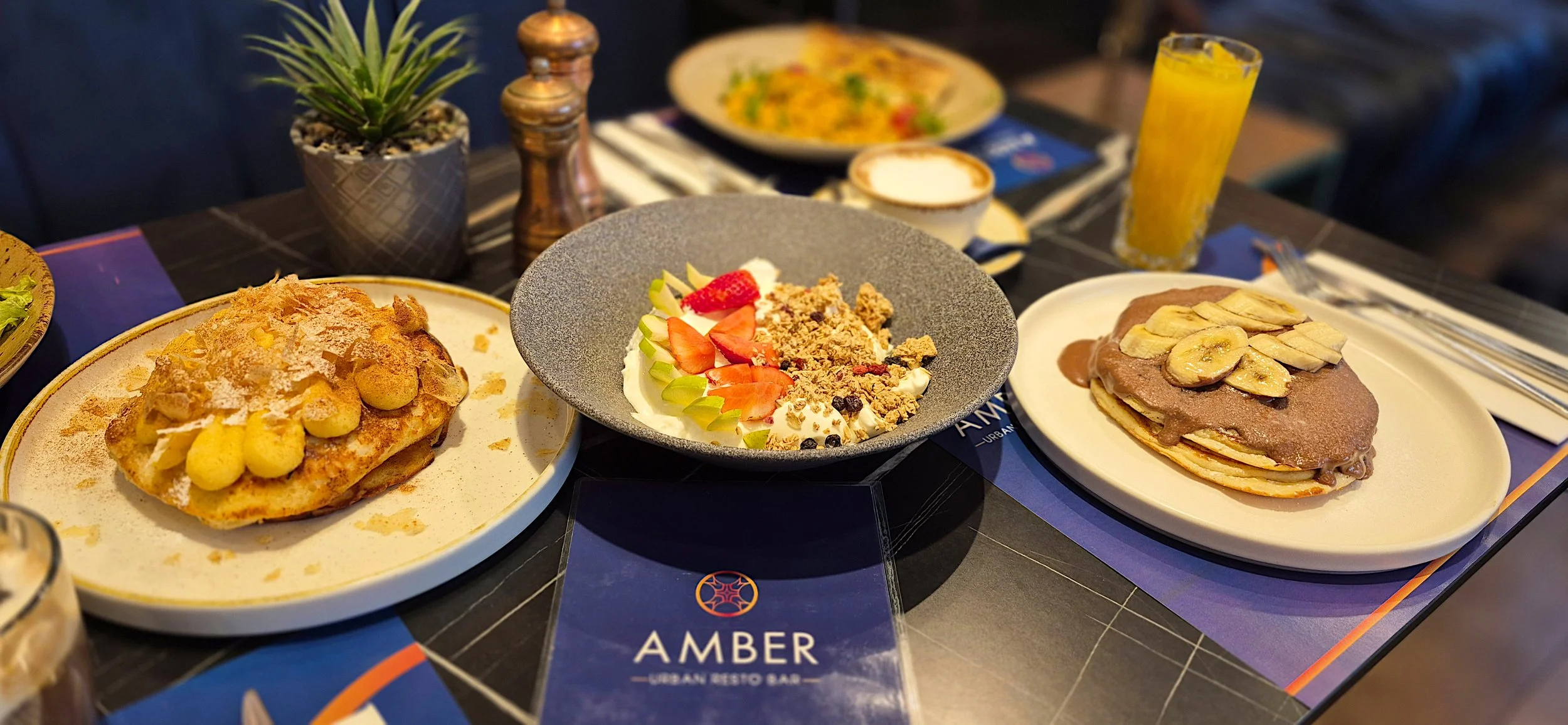 A table filled with various breakfast foods, including a plate of French toast topped with bananas and powdered sugar, a bowl of yogurt with strawberries, granola, and cucumber slices, and a stack of pancakes with banana slices and chocolate sauce. T