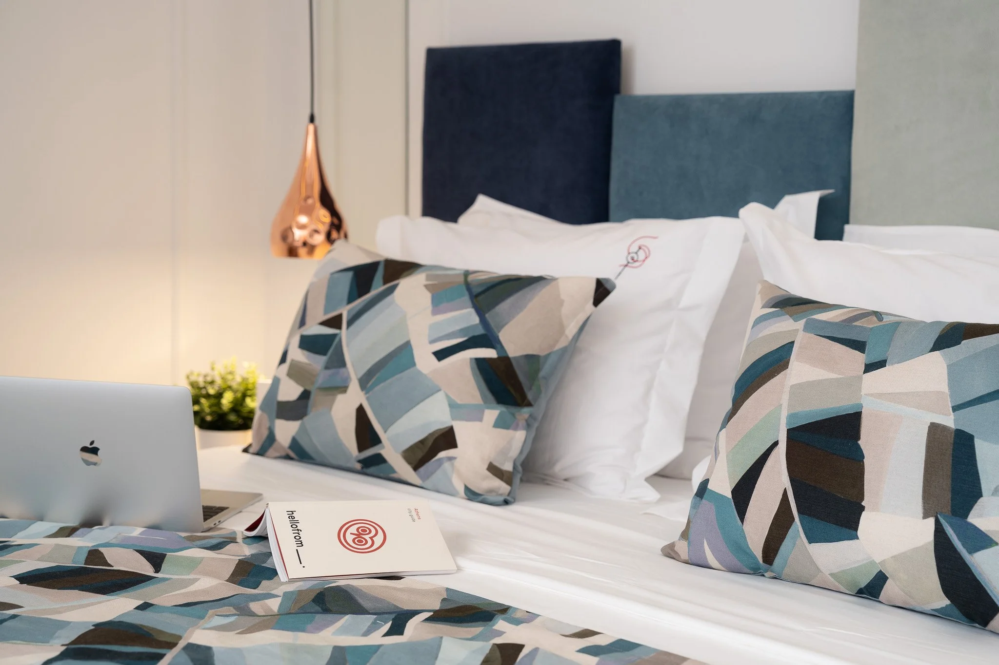 A neatly made bed with colorful geometric patterned pillows, two white pillows, a laptop, an open brochure, and a bedside lamp in a modern hotel room.