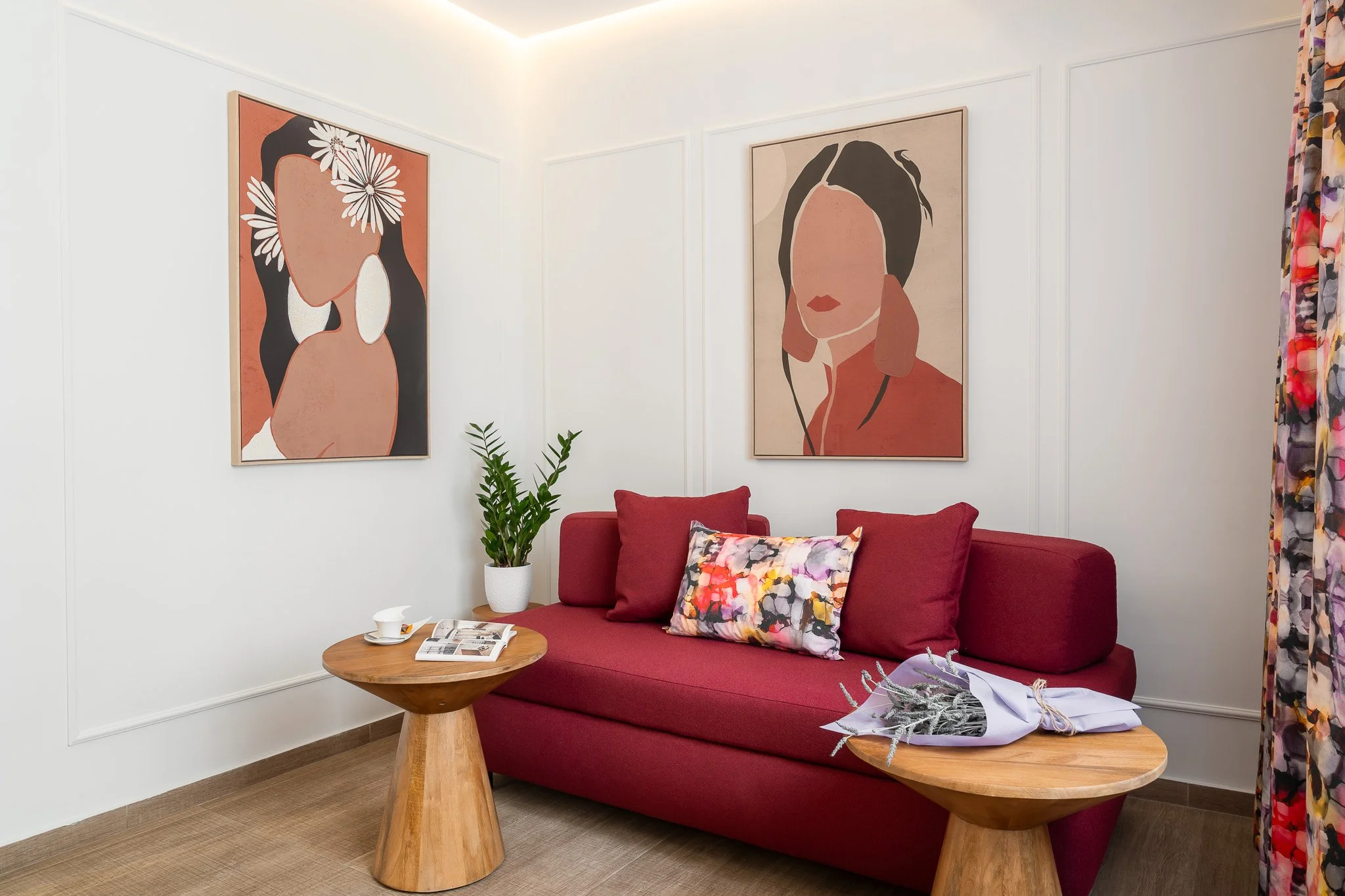 A red couch with a colorful pillow, wooden side tables, and a potted plant in a room with white walls, featuring two large abstract art portraits of women and floral curtains.