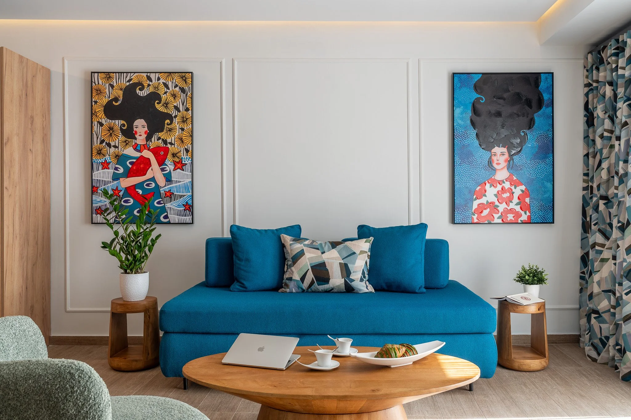 Living room with a blue sofa, two colorful paintings of women with styled hair on the wall, two small wooden side tables, and a wooden coffee table with a laptop and snacks, patterned curtains on the right.