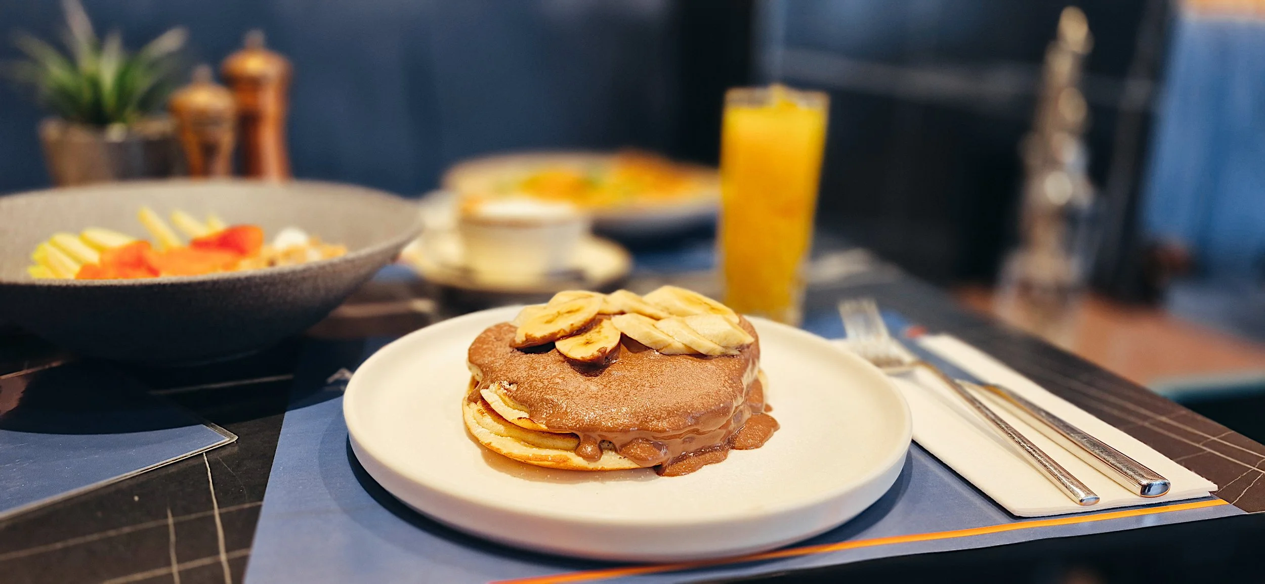 Plate with a stack of pancakes topped with sliced bananas and syrup, accompanied by a fork and knife on a napkin, set on a table with a blue placemat. In the background, there are bowls of fruit salad, a glass of orange juice, and a bowl of cereal.