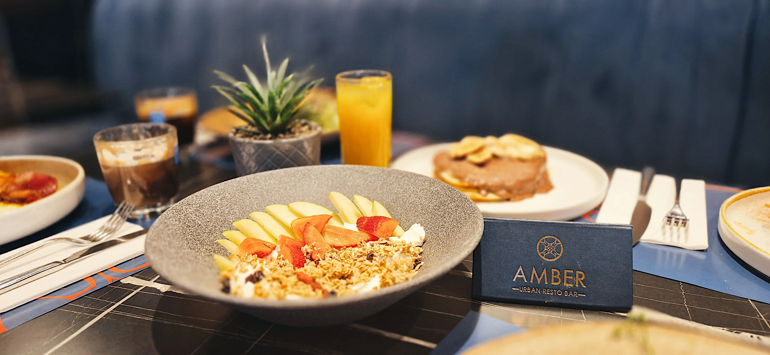 Table with assorted dishes including a fruit salad with melon and strawberries, a plate with toast topped with bananas, and a small potted plant, with drinks including orange juice and coffee cups, at Amber urban restaurant.