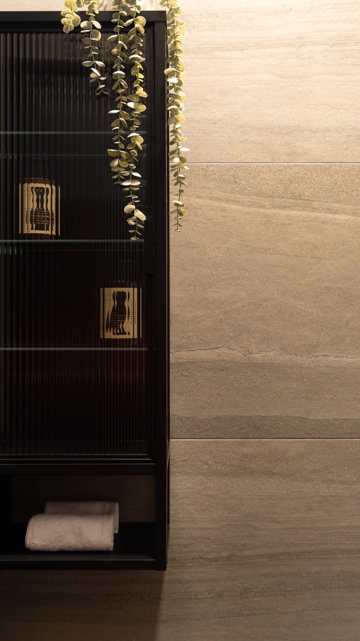 Part of a black shelving unit with rolled white towels on the bottom shelf, hanging green foliage above, and a tiled beige wall in the background.
