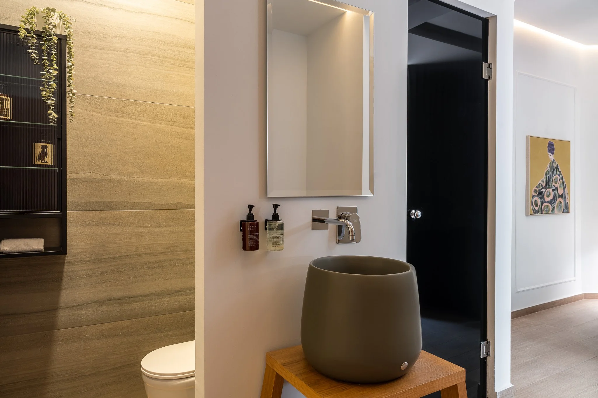 Modern bathroom with a gray vessel sink on a wooden table, a rectangular mirror above, soap and lotion dispensers, a wall vent, and a partial view into a toilet and another room with art.
