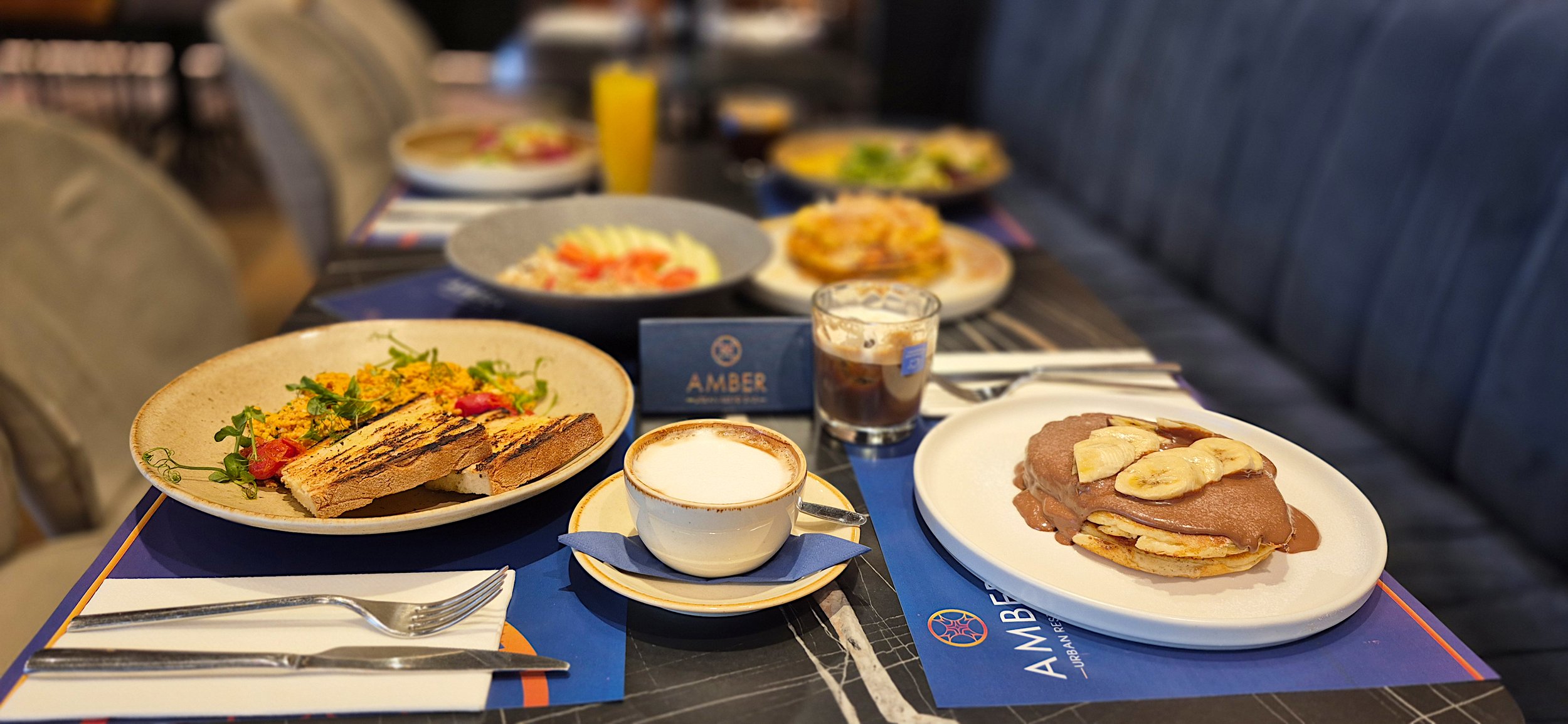 Breakfast spread on a table with plates of toast, salad, pancakes with banana and chocolate, a cup of coffee, a glass of soda, and a bottle of orange juice.
