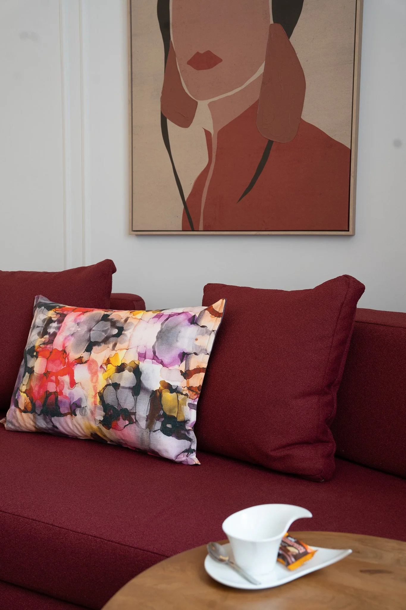 Living room with a red couch, colorful abstract pillow, framed minimalist artwork of a woman's face, white wall, wooden table with a white cup and spoon, and a sugar packet.
