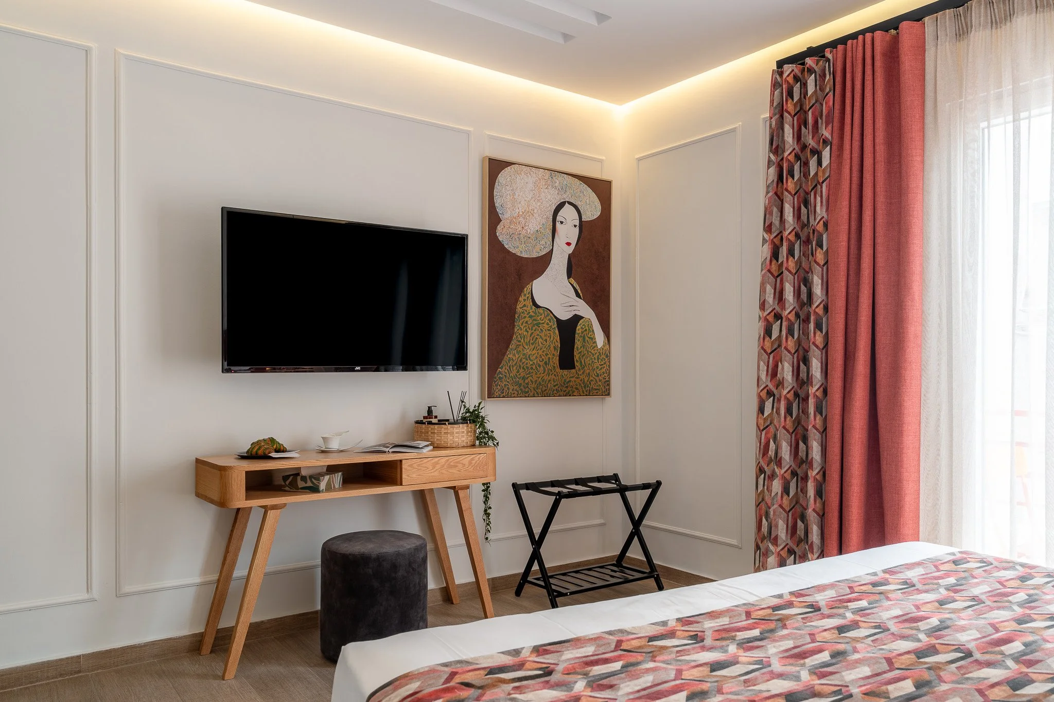 A bedroom corner featuring a wall-mounted TV, a wooden desk with a black cushioned stool, abstract art of a woman on the wall, and pink curtains beside a balcony door.