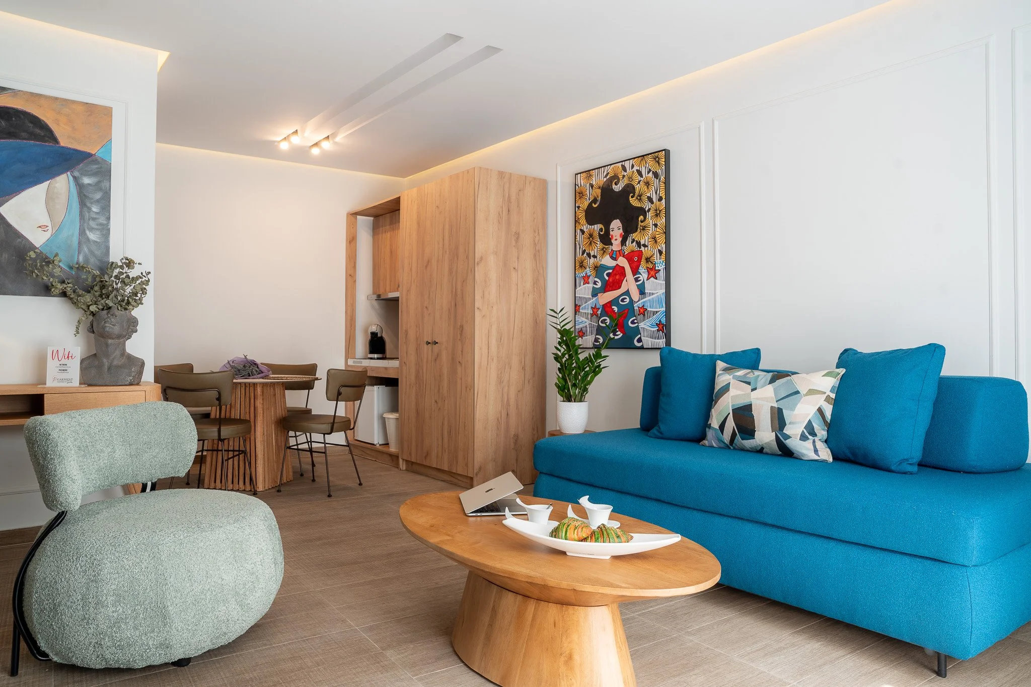 Modern living room with a blue sofa, patterned cushions, a wooden coffee table with decorative items, and a wall with colorful artwork.