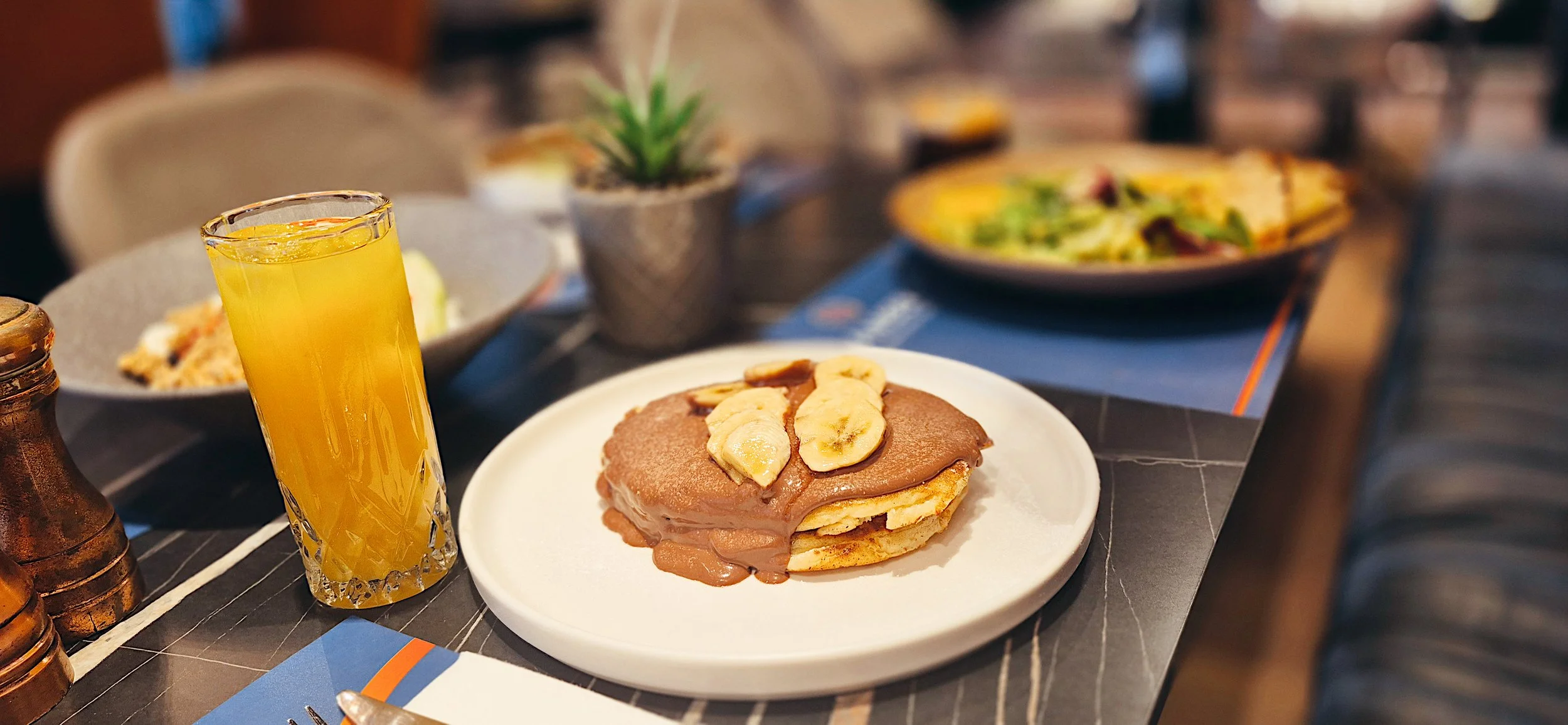 A plate with a stack of pancakes topped with banana slices and chocolate spread. Next to it is a glass of orange juice. In the background, there are bowls of food, a small potted plant, and a menu on a dark table.