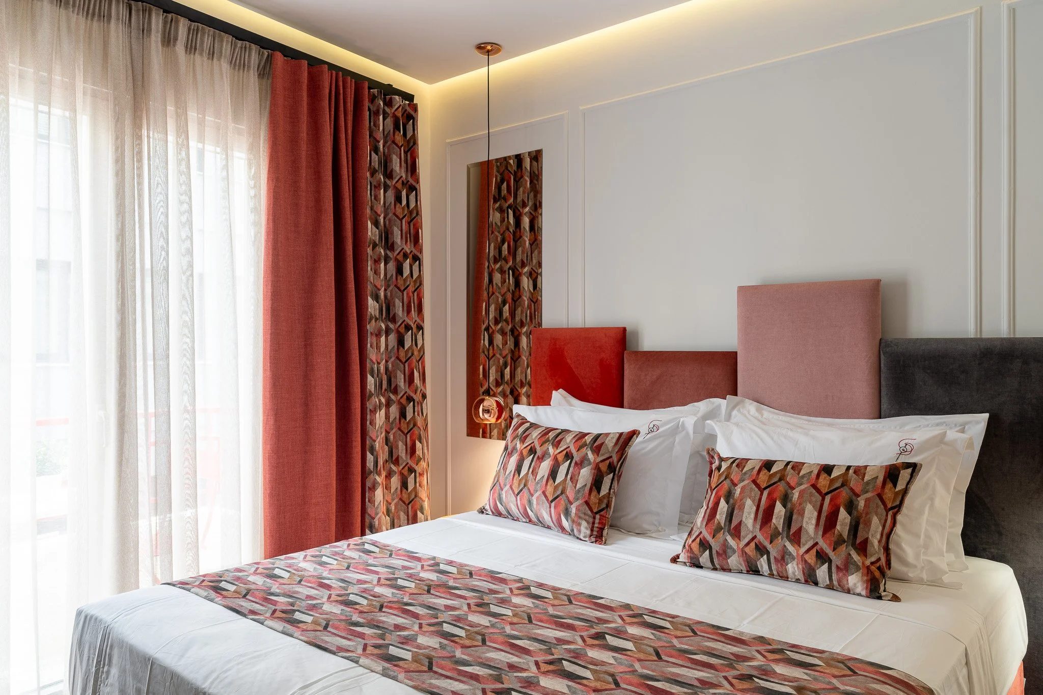 A hotel bedroom with colorful headboards, patterned pillows, and curtains, with a bedside hanging lamp.