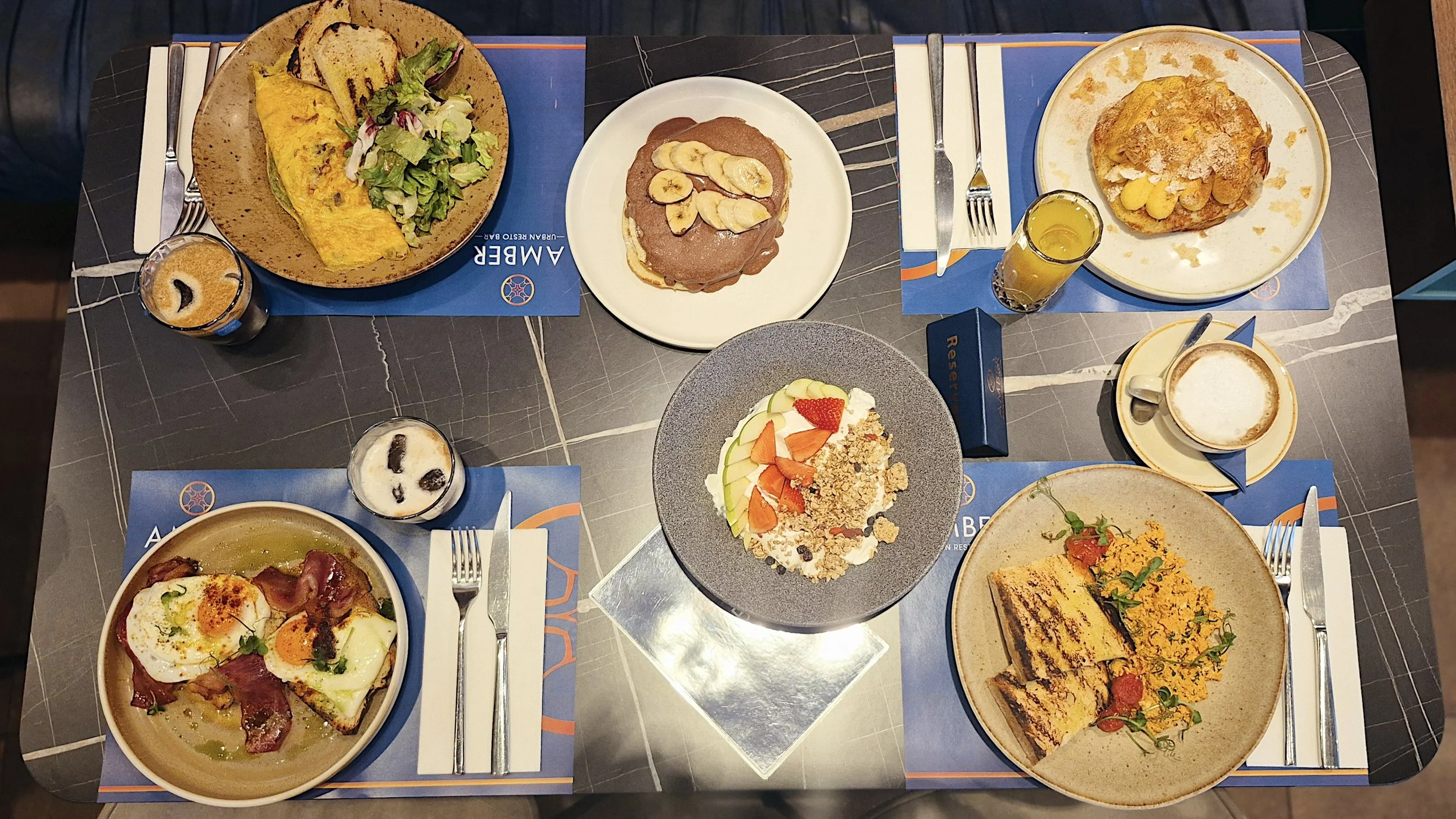 A top-down view of a breakfast or brunch table with plates of food, drinks, and desserts. Includes an omelette with toast and salad, sliced bananas on a pancake, a fruit and granola bowl, eggs with bacon, and slices of cake with yellow crumb. There a