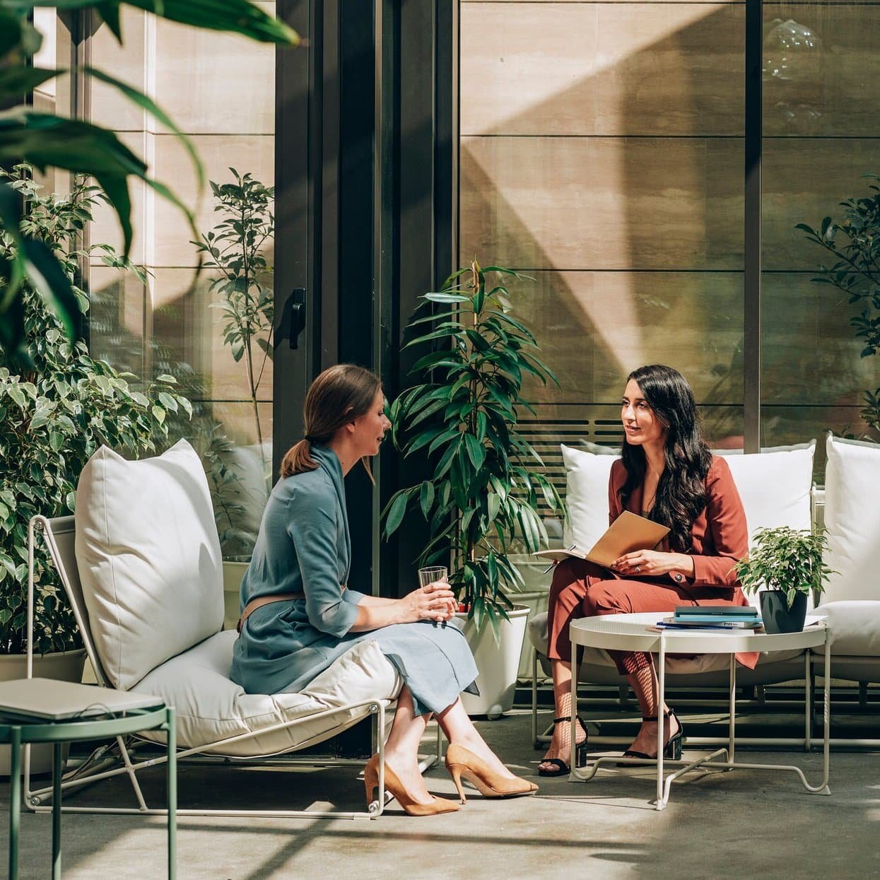 Two women having a conversation in a modern indoor lounge area with large windows, surrounded by green plants, sitting on white cushioned sofas, with a small table holding books and a potted plant. Personalised Breast Screening in Melbourne.