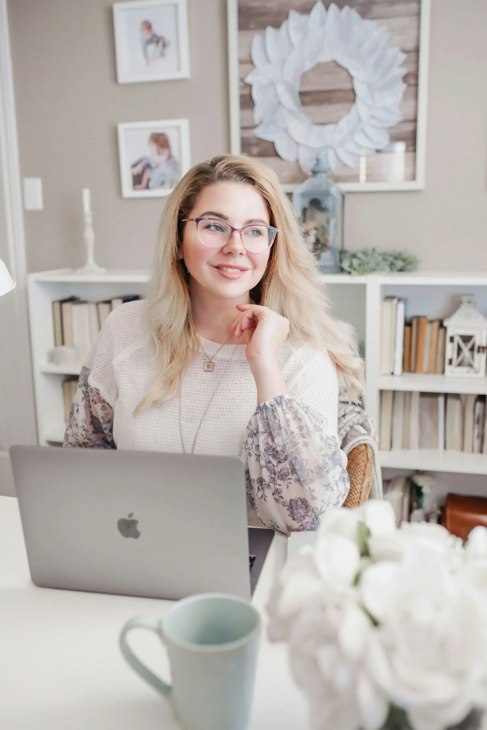 A young woman with glasses and blonde hair sitting at a desk with a silver MacBook, in a cozy, decorated room, smiling. Personalised Breast Screening is the new standard in Breast Care.
