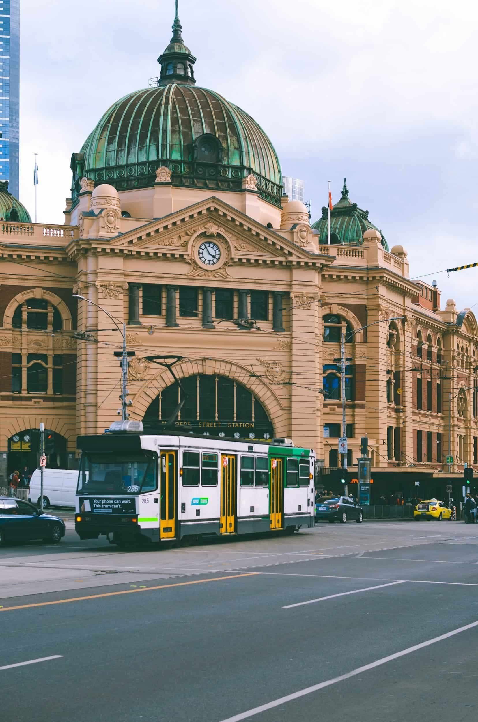 A historic train station with a domed roof and ornate architecture in an urban setting, with a tram and cars on the street in front.